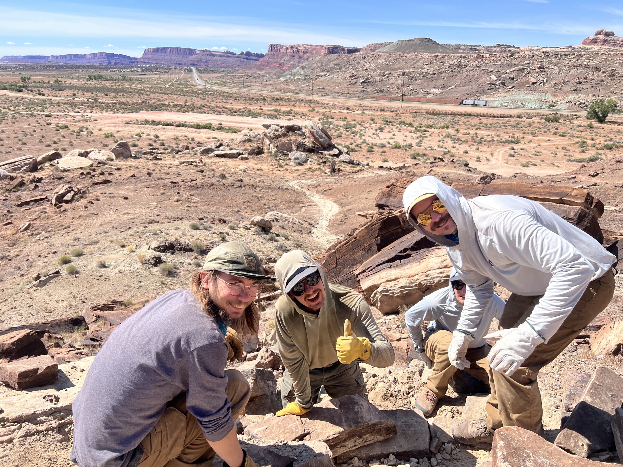 Four people in sun hats and sunglasses dig in a rocky desert area under a blue sky.